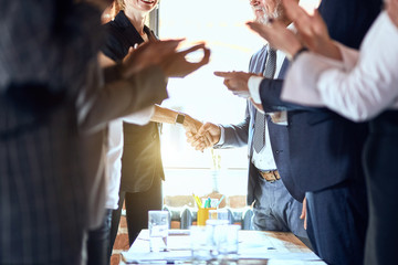 Group of businessmen at table in office. Focuced on shaking hands. Others applaud.
