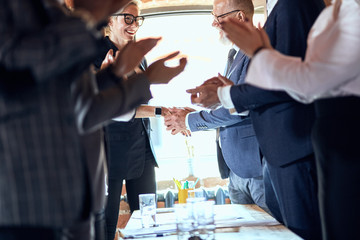 Group of businessmen at table in office. Focuced on shaking hands. Others applaud.