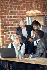 Four businessmen wear different office clothes actively discuss task at table in office. Brick wall background.