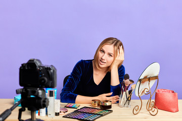 Young female beaty expert with blonde hair waiting video conference to start, talking and gazing in camera, sitting in front of table with creams, make up products, mirror. Indoor shot, isolated view.