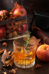Glass of alcoholic whiskey drink with a branch of rosemary with smoke and apples, cinnamon sticks. Concept of magical autumn relaxed and cozy atmosphere.
