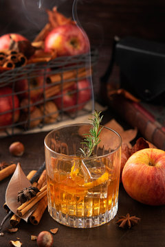 Glass of alcoholic whiskey drink with a branch of rosemary with smoke and apples, cinnamon sticks. Concept of magical autumn relaxed and cozy atmosphere.
