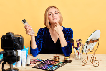 Attractive self-confident woman holding primer, describing its benefits, presenting new cosmetics line, shooting video review for blog, gazing in camera, sitting at dressing table in studio.