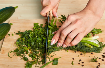 Cropped shot of womans hands chopping scallions and parsley on wooden cutting board with large knife, prepares healthy vegetarian salad for dinner. People, food and cooking concept