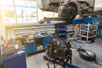 An old used car without wheel, raised on a lift for repair and under it a detached engine suspended on a blue crane near workbench in a vehicle repair workshop.