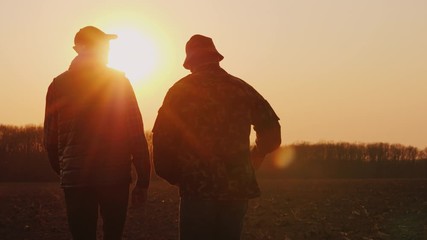 An elderly and young farmer go together over a plowed field at sunset