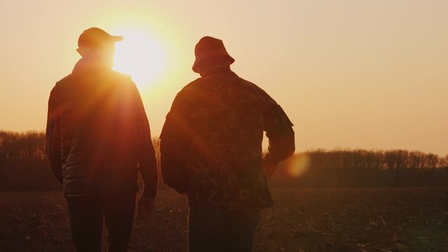 An elderly and young farmer go together over a plowed field at sunset