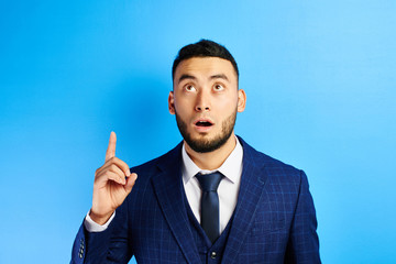 Delighted Asian Kazakh businessman has an idea, raised index finger with open mouth in a suit and tie in blue studio isolated