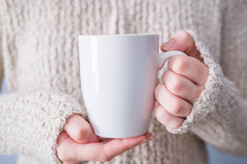 Woman in warm sweater is holding white mug in hands