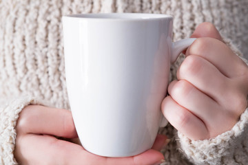 Girl in warm sweater is holding white mug in hands