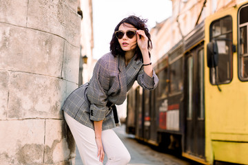 Obraz premium Portrait of a beauty brunette fashion model with sunglasses, stylish jacket and white pants. Outdoor shot. Woman in the city street, posing near the wall of building, yellow tram on the background