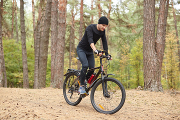 Fototapeta na wymiar Photo of cyclist on forest trail, trip on bikes in lush green nature, young attractive guy wearing black sportwear rides around trees in forest, spends free time in open air. Healthy lifestyle concept