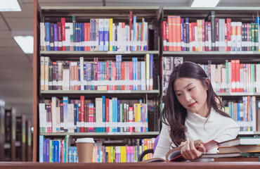 Pretty Asian female student study textbook in library with bookshelf as background in university or college. Concept of education, examination preparation, reading, learning and knowledge searching