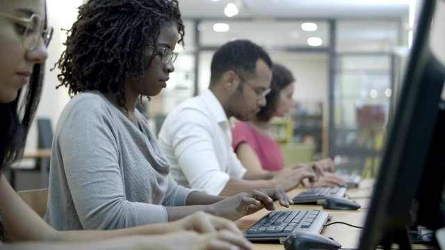 Side view of people sitting at tables and working with computers. Row of workers typing on keyboards of computers. Technology concept