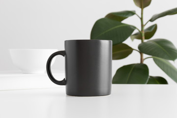Black mug mockup with workspace accessories on a white table and a ficus plant.