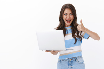 Fototapeta premium Lively satisfied modern gorgeous woman picked cool computer, smiling broadly hold laptop, show thumb up in approval, accept gesture, like cool friend project music-video, leave positive comment