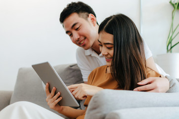 young asian sweet love couple talking and using mobile tablet, watching movie online on sofa at home office, internet technology, social media, online booking, entertainment, shopping online concept