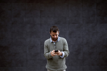Serious bearded fashionable Caucasian man in sweater standing outdoor and using smart phone for typing message.