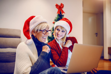 Happy senior woman and her daughter sitting on floor in living room and using laptop for christmas shopping. Both having santa hats on heads. In background is christmas tree.