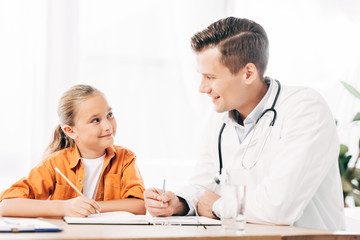 smiling child and pediatrist looking at each other while writing in notebook at table in clinic