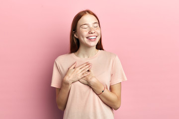 friendly positive woman smiles sincerely, keeps both palms on heart, dressed in casual pink t...