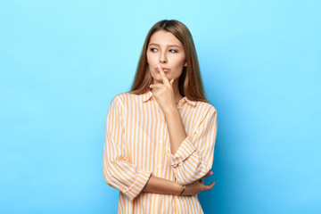 Thoughtful serious beautiful woman with long brown hair keeps finger near lips, being deep in thoughts,looking aside, thinks about coming holidays, isolated on blue background . copy space