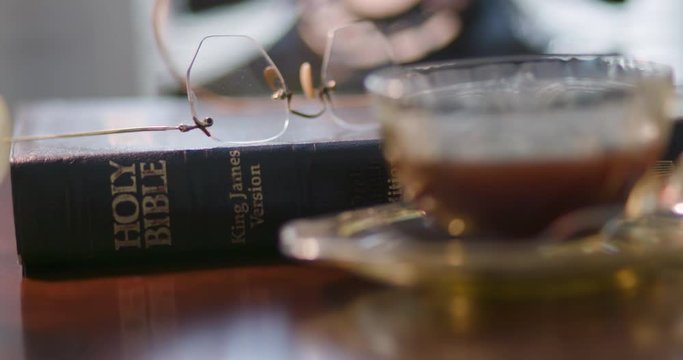 Scene Tracks Across The Surface Of A Desk Where Depression Period Glass Coffee Cup With Matching Creamer Are Placed In The Foreground And A Closed Bible With Vintage Eyeglasses Laying On Top In The Ba