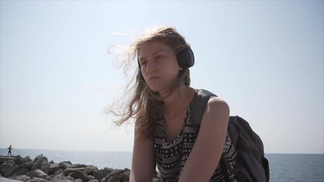 A girl with a backpack and headphones listens to music while sitting on the promenade on a sunny day. Slow motion