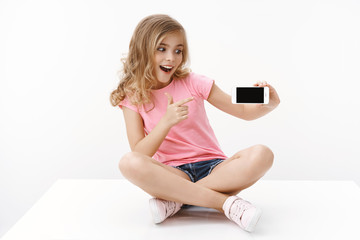 Cheerful excited cute blond little girl sitting on floor with crossed legs, hold smartphone,...