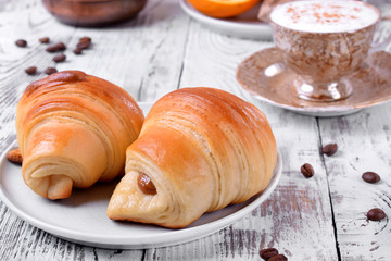 Two croissants of yeast dough on ceramic plate, freshly brewed coffee in a cup and oranges on white wooden table. Breakfast idea