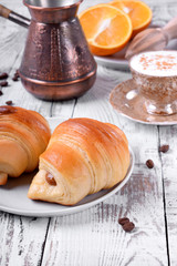 Two croissants of yeast dough on ceramic plate, freshly brewed coffee in a cup and oranges on white wooden table. Breakfast idea