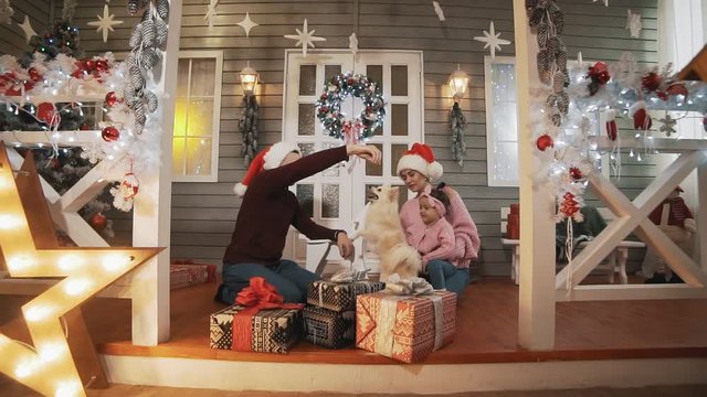 Young family with little daughter and dog sitting on porch with Christmas tree and decor. Happy family concept in slow motion. Young family in pullovers hugging in Christmas day