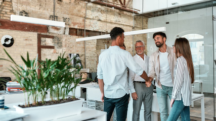 Welcome! Two men shaking hands and smiling while standing with coworkers in the creative office
