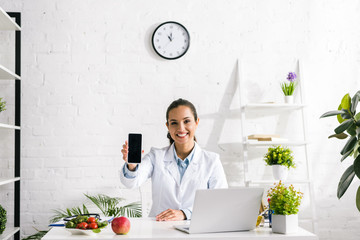 happy nutritionist holding smartphone with blank screen near laptop