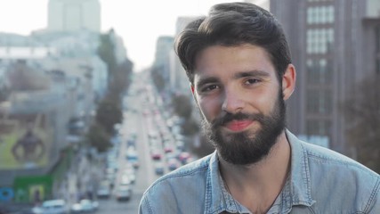 Young bearded man smiling to the camera with city on background. Handsome male hipster relaxing outdoors, looking to the camera joyfully. Youth, lifestyle concept