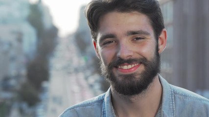 Cheerful handsome bearded man smiling to the camera. Cropped portrait of a charming young man looking to the camera with a smile, busy city streets on background