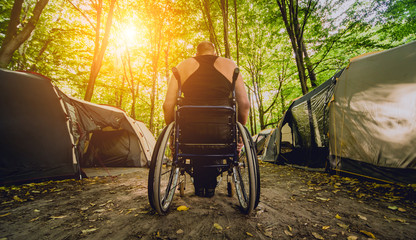 Disabled man resting in a campsite with friends. Wheelchair in the forest on the background of tents