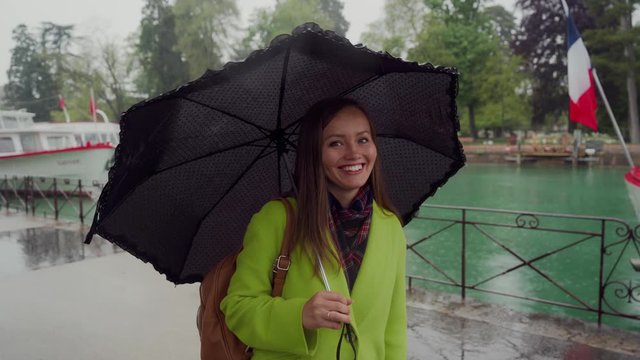 Beautiful girl in a yellow coat stands in the rain, twists a cute black umbrella, smiles and flirts. Lake Annecy, France. Portrait