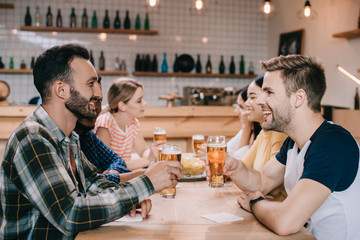 cheerful multicultural friends talking and drinking light beer in pub