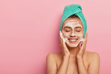 Pleased smiling woman washes face with cleansing gel, has soap on complexion, keeps eyes shut,...