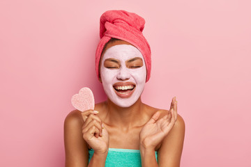 Portrait of overjoyed woman laughs happily, holds cosmetic sponge, raises palm being in high...
