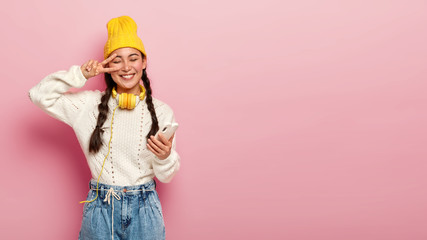 Positive lovely Asian lady makes peace gesture over eye, wears yellow hat, stylish sweater with jeans, uses mobile phone for downloading songs in playlist, headphones connected to some device