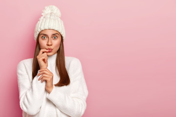 Horizontal shot of surprised dark haired woman looks surprisingly, keeps lips rounded, wears snow white winter hat and sweater, dressed in warm outfit poses against pink background. Fashion and season