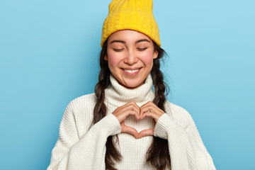 Indoor shot of beautiful brunette woman keeps eyes closed with pleasure, makes heart gesture,...