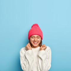 Vertical image of joyful young girl feels overjoyed, raises clenched fists, being in good mood, wears white sweater and pink hat, dressed in warm clothes during cold autumn day, isolated on blue wall
