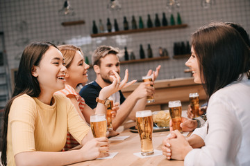 cheerful multicultural friends talking while drinking beer together in pub