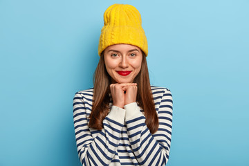 Image of tender good looking dark haired woman stands against blue background in yellow hat and striped sweater, wears red lipstick, makeup, looks staightly at camera. People, beauty, fashion - obrazy, fototapety, plakaty