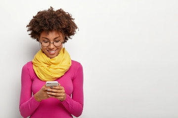 Horizontal shot of pleased dark skinned female holds modern mobile phone, focused into display,...