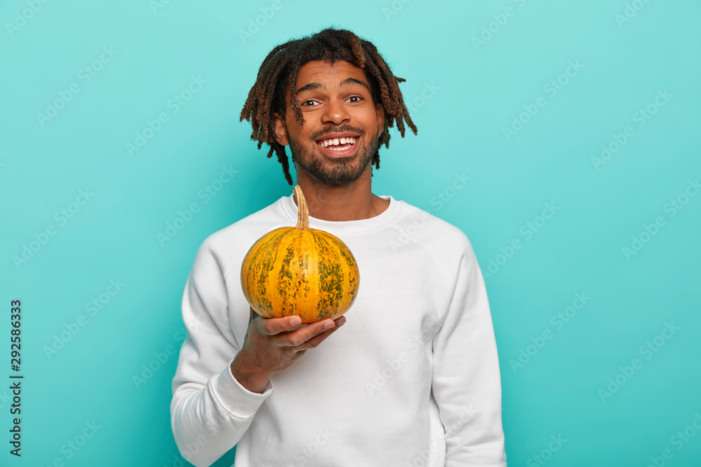 Wall mural Happy handsome young mixed race guy with dreads, holds little ripe yellow pumpkin, going to make diet salad, dressed in casual wear, carves vegetable for Halloween, isolated over blue background. - Wall murals