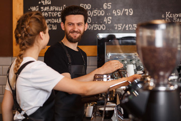 Two young smiling barista at work. Professional barista team brewing coffee using coffee machine in coffee shop. Happy young man and woman developing own coffee business. Coffee shop concept.